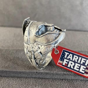 Puede incluir: Un anillo plateado con un diseño detallado y texturizado. El anillo tiene una apariencia esculpida única. Una etiqueta roja con el texto "TARIFF FREE" está adjunta al anillo.