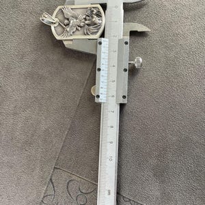 May include: A silver pendant with a detailed design of an angel with wings spread. The pendant is being measured with a ruler, showing it is approximately 3.5 cm in length.