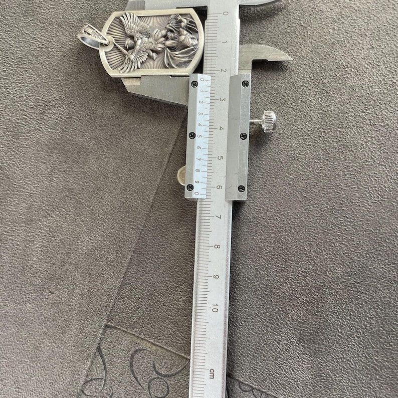 May include: A silver pendant with a detailed design of an angel with wings spread. The pendant is being measured with a ruler, showing it is approximately 3.5 cm in length.