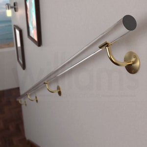 Puede incluir: Un pasamanos de acrílico transparente con soportes de metal dorado montados en una pared blanca. El pasamanos es una adición moderna y elegante a cualquier hogar.