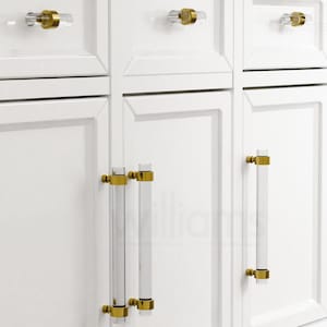 Könnte beinhalten: Weiße Schranktüren mit goldenen und durchsichtigen Glas-Schrankgriffen. Die Griffe sind rechteckig und haben eine zylindrische Form.