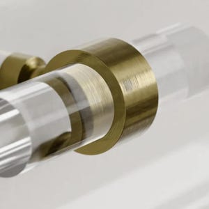 Puede incluir: Un pomo de armario de vidrio transparente y dorado con forma cilíndrica. El pomo tiene un acabado dorado cepillado y un cilindro de vidrio transparente con textura acanalada.