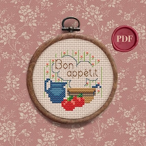 Peut inclure: Un cerceau de broderie au point de croix avec un pichet bleu, un panier de fruits et le texte "Bon appétit" en lettres vertes et rouges.