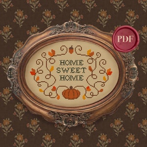 May include: A cross-stitch pattern featuring a pumpkin and the text "Home Sweet Home" surrounded by autumn leaves and vines. The pattern is framed in a brown oval frame.