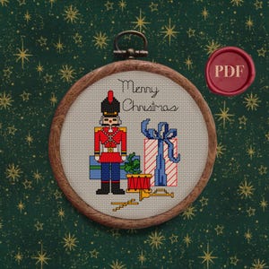 May include: A cross-stitch pattern featuring a toy soldier holding a drum and a gift box with a bow. The pattern is framed in a wooden hoop and includes the text "Merry Christmas".