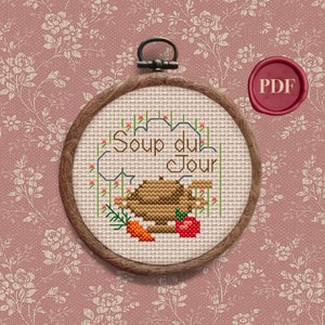 May include: A cross-stitch embroidery hoop with a brown wooden frame. The embroidery depicts a brown teapot with a carrot and an apple, and the text "Soup du Jour".