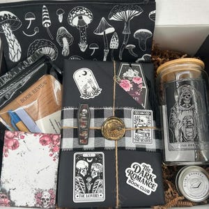 May include: A gift box containing a black zippered pouch with white mushroom illustrations, a book review notepad, a black wrapped book with tarot-themed stickers and a wax seal, a glass tumbler with a skeleton graphic, and a small round tin.