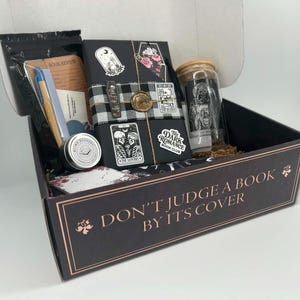 May include: A black gift box with the text DON'T JUDGE A BOOK BY ITS COVER containing a wrapped book with tarot-style stickers, a glass tumbler with a skull graphic, a blue pen, a small round tin labeled BLIND BOOKERY, and stationery items.