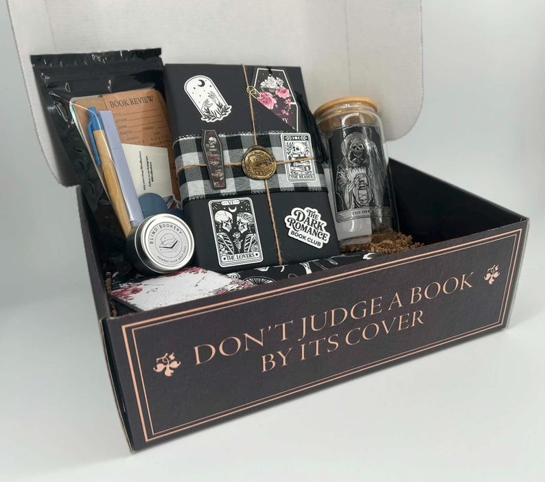 May include: A black gift box with the text DON'T JUDGE A BOOK BY ITS COVER containing a wrapped book with tarot-style stickers, a glass tumbler with a skull graphic, a blue pen, a small round tin labeled BLIND BOOKERY, and stationery items.