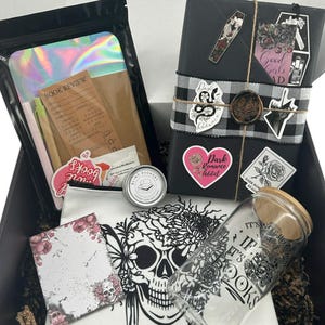 May include: A gift box containing a black-wrapped book with stickers and a wax seal, a holographic pouch with a book review card, a small round tin, a white canvas bag with a skull print, and a glass tumbler with a bamboo lid and book-themed text.