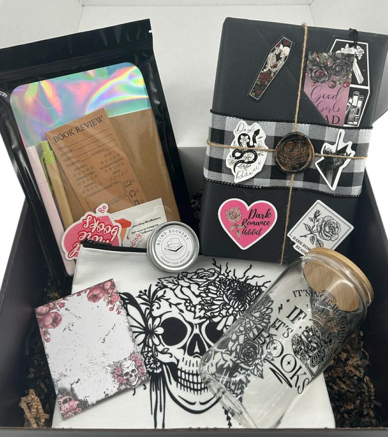 May include: A gift box containing a black-wrapped book with stickers and a wax seal, a holographic pouch with a book review card, a small round tin, a white canvas bag with a skull print, and a glass tumbler with a bamboo lid and book-themed text.