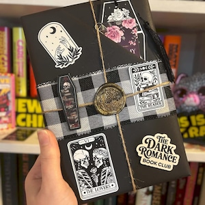 May include: A black wrapped book decorated with a black and white buffalo check ribbon, a gold wax seal, and twine. Stickers include a moon and hands, a coffin with a skull, a reader card, a lovers tarot card, and a Dark Romance Book Club label.