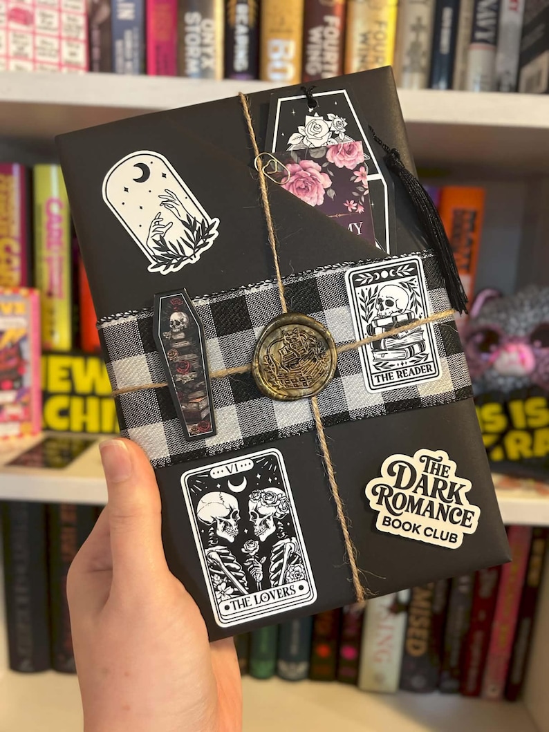 May include: A black wrapped book decorated with a black and white buffalo check ribbon, a gold wax seal, and twine. Stickers include a moon and hands, a coffin with a skull, a reader card, a lovers tarot card, and a Dark Romance Book Club label.