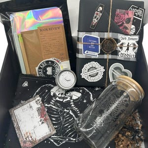May include: A gift box containing a book wrapped in black paper with stickers and a wax seal, a glass tumbler with a bamboo lid, a black zippered pouch with a skeleton print, a book review notebook, a small metal tin, and a holographic pouch.