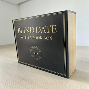 May include: A black book box with gold lettering that reads "BLIND DATE WITH A BOOK BOX." The box has a gold border and a logo that says "Blind Bookery." The side of the box has a light brown, wood-like appearance.