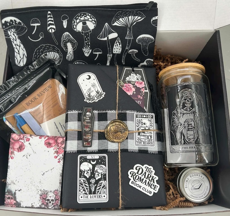 May include: A gift box containing a black zippered pouch with white mushroom illustrations, a book review notepad, a black wrapped book with tarot-themed stickers and a wax seal, a glass tumbler with a skeleton graphic, and a small round tin.