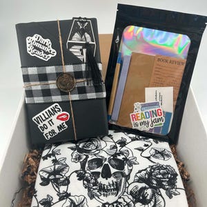 May include: A gift box containing a black-wrapped book with a buffalo plaid ribbon, a wax seal, and stickers reading Dark Romance Reader and VILLAINS DO IT FOR ME. Includes a holographic pouch with stationery and a white cloth featuring a black skull print.