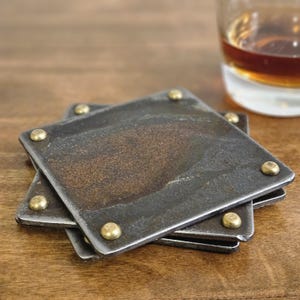 May include: A stack of three square, rustic metal coasters with rounded brass rivets in each corner. The coasters have a dark, weathered finish and are stacked on a wooden surface. A glass of amber liquid is in the background.