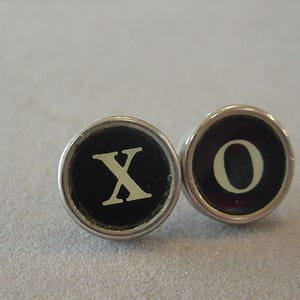 May include: Two circular typewriter keys featuring a black background with a silver-toned metal rim. The left key displays the white letter X, and the right key displays the white letter O. These vintage-style keys are positioned side by side.