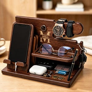 May include: A dark brown wooden desk organizer featuring a phone stand, a watch holder, a shelf for glasses, and compartments for small items. The wood is engraved with the name Miko and the initials MVI. It holds a smartphone, watch, glasses, pen, and keys.