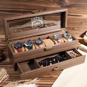 May include: A wooden watch box with a glass top and a drawer. The box has a brown finish and is engraved with the text "SELECTION 1982 VINTAGE". The box has six slots for watches and a drawer for storing other items.