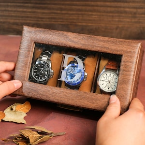 Könnte beinhalten: Eine Holzuhrbox mit Glasdeckel, in der drei Uhren mit unterschiedlichen Stilen und Farben ausgestellt sind. Die Box hat eine personalisierte gravierte Initial auf der Vorderseite.