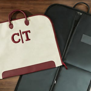 Puede incluir: Funda de ropa color crema con detalles y asas burdeos, con las iniciales "C|T" bordadas. Una funda de ropa negra está abierta al lado, mostrando un bolsillo transparente. Las fundas están sobre una superficie de madera.