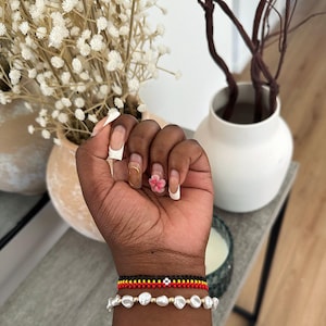 May include: A wrist adorned with three bracelets: a strand of white pearls, a black and red beaded bracelet, and a red and yellow beaded bracelet. The hand displays manicured nails with a floral accent and gold and white designs. A vase with dried flowers is in the background.