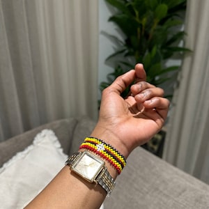 Op de afbeelding: Een zilveren en gouden horloge met een vierkante wijzerplaat en een kralenarmband in de kleuren van de vlag van Oeganda.
