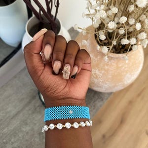 May include: A person's wrist wearing two bracelets. The top bracelet is turquoise blue and has a small white diamond in the center. The bottom bracelet is made of white pearls.