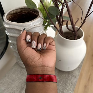May include: A red beaded bracelet with a small green heart design. The bracelet is on a person's wrist.