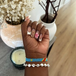 Könnte beinhalten: Eine Hand mit weißem und rosafarbenem Nagellack und einem Blumenring am Ringfinger. Die Hand trägt ein blaues Perlenarmband mit gelben und roten Akzenten und ein weißes Perlenarmband.