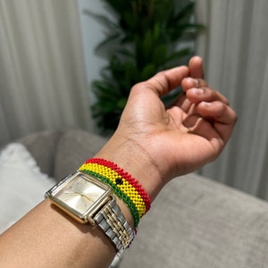 Puede incluir: Un reloj dorado con una esfera cuadrada y una correa de metal plateado y dorado. El reloj se lleva en la muñeca de una persona con una pulsera de cuentas rojas, amarillas y verdes.