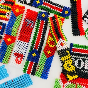 Op de afbeelding: Een verzameling kleurrijke kralenarmbanden, elk met de vlag van een ander land. De armbanden zijn gemaakt van kleine, ronde kralen in verschillende kleuren, waaronder rood, wit, blauw, groen, geel en zwart. De armbanden zijn willekeurig gerangschikt op een wit oppervlak.