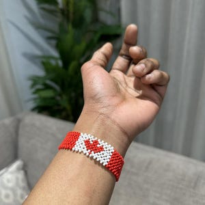 May include: A red and white beaded bracelet with a red maple leaf design. The bracelet is worn on a person's wrist.
