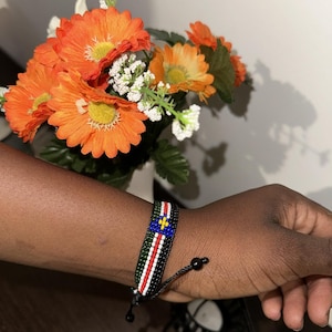Può includere: Un braccialetto di perline nere con un design colorato che ricorda la bandiera di Saint Kitts e Nevis. Il braccialetto ha un cordoncino nero con un perno all'estremità per regolare la vestibilità.