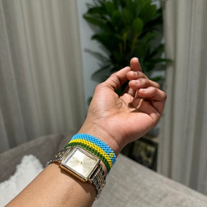 Può includere: Un orologio quadrato dorato con un cinturino in metallo argentato al polso di una persona. L'orologio è indossato con un braccialetto di perline nei colori della bandiera panafricana: verde, giallo e blu.