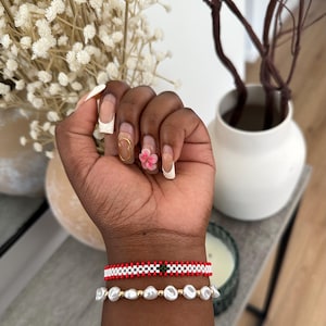 Könnte beinhalten: Zwei Armbänder an einem Handgelenk. Das obere Armband ist rot, weiß und grün mit einem kleinen grünen Quadrat in der Mitte. Das untere Armband ist weiß mit perlmuttartigen Perlen.