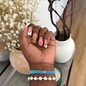 May include: A person's wrist with two bracelets. The top bracelet is turquoise blue with a small white flower charm. The bottom bracelet is white with pearl-like beads.