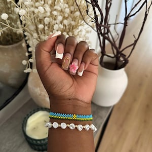 Può includere: Un polso mostra due braccialetti. Il braccialetto superiore è realizzato con perline blu, gialle e verdi. Il braccialetto inferiore presenta perle bianche di forma irregolare. La mano ha una manicure francese con un anello d'oro e un fiore rosa.