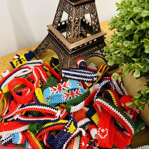 May include: A collection of colourful beaded bracelets, each displaying a different country's flag design. The bracelets are arranged around a miniature Eiffel Tower, suggesting a theme of travel or international friendship.