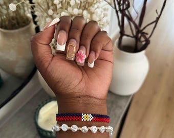 Puede incluir: Una muñeca con dos pulseras. La pulsera superior es roja, azul y blanca con un diseño de flor amarilla. La pulsera inferior es blanca y está hecha de perlas.