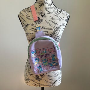 May include: A purple and green Stitch backpack with a pink adjustable strap. The backpack features a patchwork design with images of Stitch in various poses. The text "Stitch" and "Ohana" are printed on the backpack.