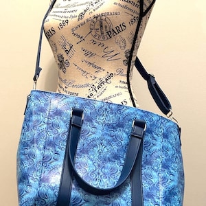 May include: A blue patterned tote bag with a matching shoulder strap and handles. The bag features a swirling design in shades of blue and is displayed on a mannequin. The bag is designed for carrying items.