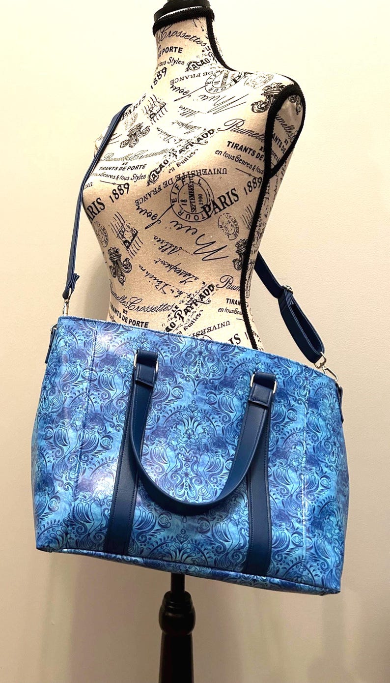 May include: A blue patterned tote bag with a matching shoulder strap and handles. The bag features a swirling design in shades of blue and is displayed on a mannequin. The bag is designed for carrying items.