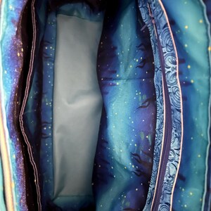 May include: Interior view of a tote bag with a celestial design. The bag's lining features a blue and teal starry night pattern with a light blue pocket and a zippered compartment with a paisley design.