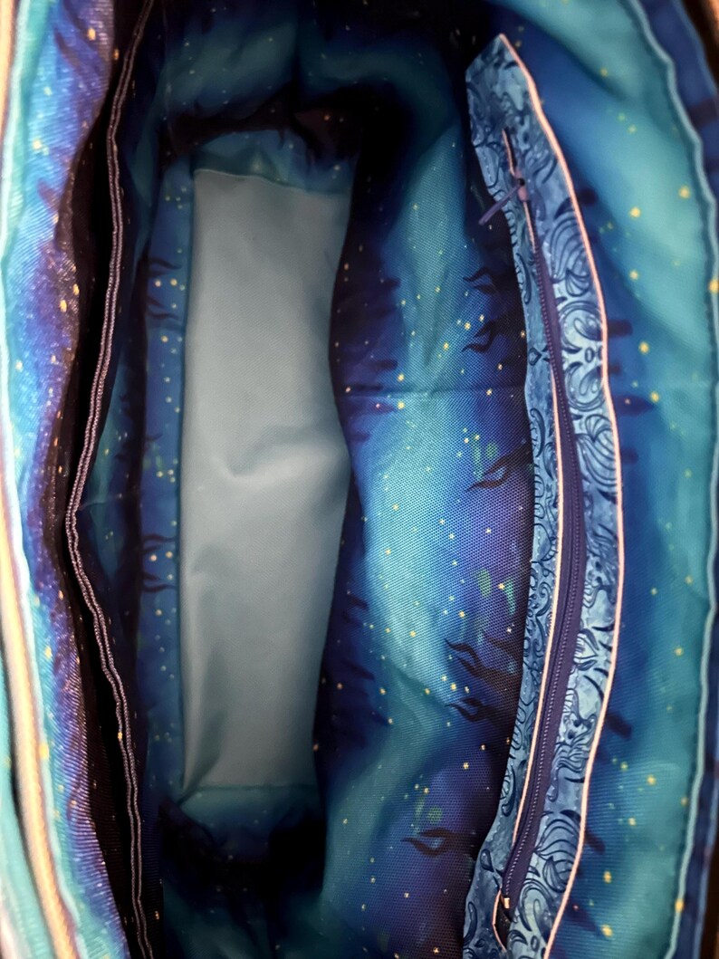 May include: Interior view of a tote bag with a celestial design. The bag's lining features a blue and teal starry night pattern with a light blue pocket and a zippered compartment with a paisley design.
