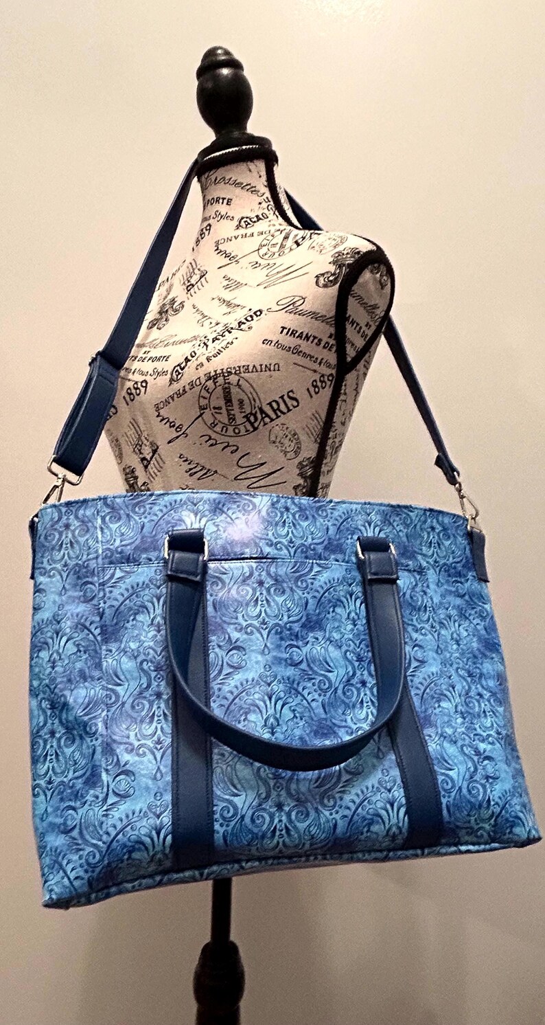 May include: Blue tote bag with a floral pattern and dark blue handles and shoulder strap. The bag is displayed on a mannequin with text that says "Paris 1889".