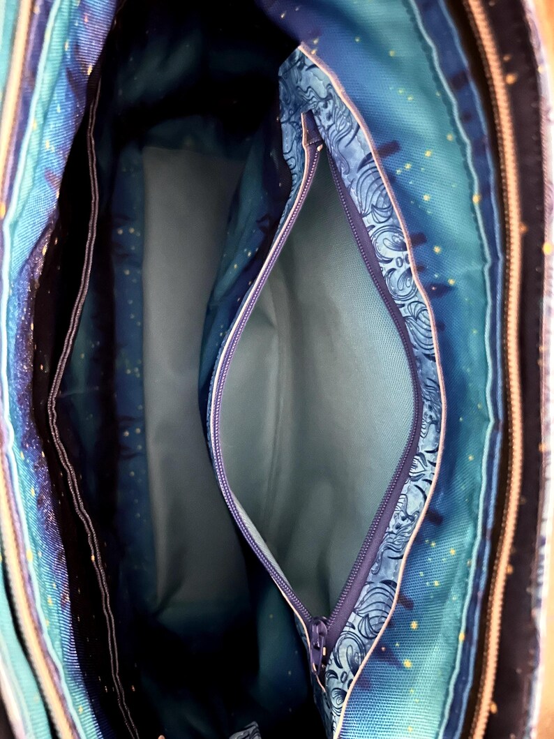 May include: Interior view of a tote bag with multiple compartments. The bag features a blue and teal patterned fabric with a zipper closure. The interior lining is a solid light blue color. The bag has a celestial theme with stars.