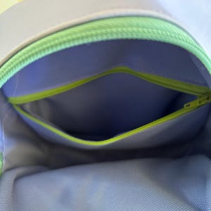 May include: A lavender-coloured bag with a green zipper and a light blue mesh lining. The bag has a small pocket on the inside.